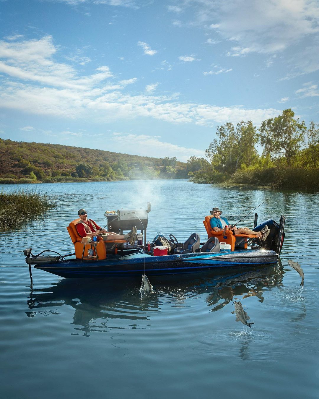 Two adult men fish and cook from a boat on a river, surrounded by trees and a blue sky. Fish jump from the water around the boat.
